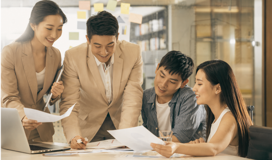 Group of four people in a modern office setting, discussing documents around a laptop.
